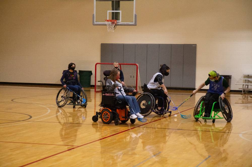 Opposing adult teams reaching for the ball with hockey sticks during a wheelchair floor hockey game.
